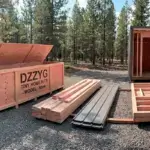 Dzzyg Tiny Home Kit Model Nook wooden crate with pre-cut building materials and assembled frame structure on gravel lot in forest setting