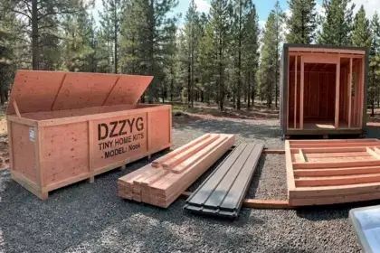 Dzzyg Tiny Home Kit Model Nook wooden crate with pre-cut building materials and assembled frame structure on gravel lot in forest setting